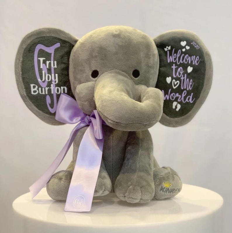 May include: A gray plush elephant with a purple ribbon and text on its ears. One ear reads "Tru Joy Burton" in purple script, the other reads "Welcome to the World" with hearts and the year 2020. The elephant has a small gold crown logo on its foot.