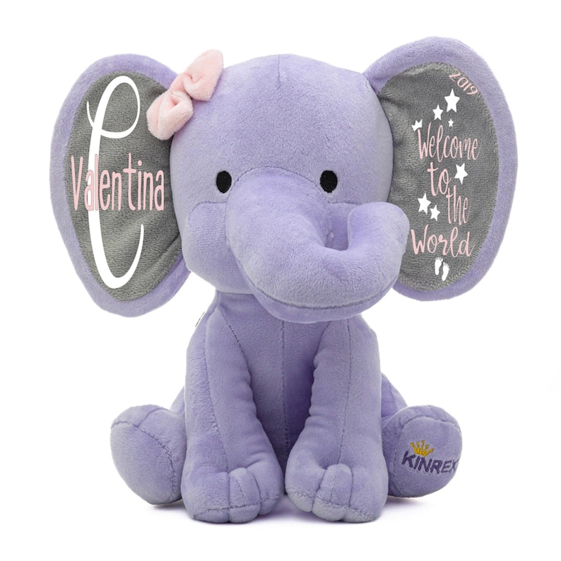 May include: A lavender plush elephant toy with a pink bow and gray ear accents. One ear reads "Valentina" and the other reads "Welcome to the World" with stars. The elephant has the word "KINREX" and a crown on its foot.