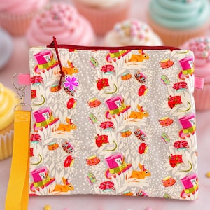 Könnte beinhalten: Eine rechteckige, gemusterte Tasche mit rotem Reißverschluss und gelber Handschlaufe. Die Tasche hat ein sich wiederholendes Design eines Hasen mit Zylinder, Teetassen und anderen skurrilen Elementen auf grauem Hintergrund. Rosa Cupcakes sind im Hintergrund zu sehen.