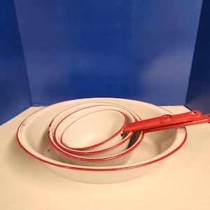 May include: Set of four white enamel bowls with red rims. The bowls are nested inside each other, with the largest bowl at the bottom and the smallest bowl at the top. The bowls are all in good condition and have a vintage look.