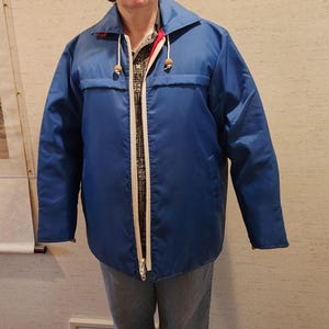 May include: A blue nylon jacket with a white zipper and a drawstring hood. The jacket has two front pockets and wooden toggles on the hood. The jacket is worn over a patterned shirt and jeans.