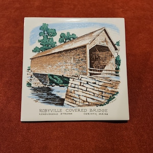 May include: A vintage tile featuring a hand-painted illustration of the Robyville Covered Bridge in Corinth, Maine. The bridge is brown with a wooden roof and is situated over a stream. The text "ROBYVILLE COVERED BRIDGE" is printed below the image, along with "KENDUSKEAG STREAM" and "CORINTH, MAINE".
