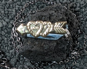 Puede incluir: Un collar dorado con un colgante que presenta dos corazones entrelazados con la palabra "LOVE" inscrita en uno de los corazones y una flecha que los atraviesa. El collar está hecho de una cadena de plata.