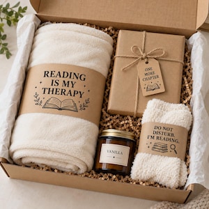 May include: A gift box containing a cream-colored fleece blanket with a band reading READING IS MY THERAPY, a brown paper-wrapped gift with a tag reading ONE MORE CHAPTER, a vanilla candle in a glass jar, and cream socks with a band reading DO NOT DISTURB. I'M READING.