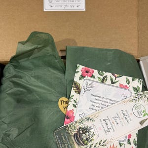 Peut inclure: Une bo&icirc;te en carton ouverte avec du papier de soie vert et des cartes. Les cartes pr&eacute;sentent des motifs floraux et le texte "Mystery Book Gift Box". Une carte dit "Joyeux anniversaires aux amoureux des livres".