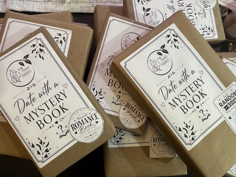 Peut inclure: Plusieurs livres emball&eacute;s avec des &eacute;tiquettes blanches indiquant "Date with a Mystery Book" et "Romance Book". Les livres sont envelopp&eacute;s dans du papier brun. Les &eacute;tiquettes pr&eacute;sentent un motif floral et les mots "The Foliage Goddess".