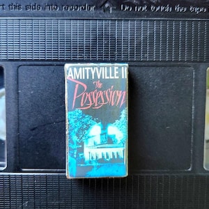 May include: A VHS tape of the movie "Amityville II: The Possession" with a blue and black cover. The title is in red and the image shows a white house with blue trees. The tape is on a black VHS cassette.