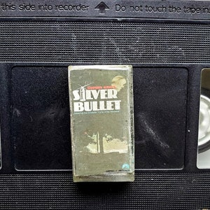 Puede incluir: Una cinta VHS vintage de la película "Silver Bullet" basada en la novela "Cycle of the Werewolf" de Stephen King. La cinta está en una caja VHS negra, con el título en blanco y el nombre del autor en la parte superior.