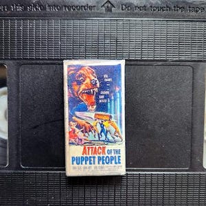 May include: A vintage VHS tape featuring the movie "Attack of the Puppet People." The cover art displays a snarling bear, figures, and the film's title in bold letters. The tape is set against a black background.
