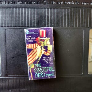 May include: A vintage VHS tape of "The Grateful Dead Movie." The cover features a colorful illustration of a skeleton in a top hat and striped suit. Text includes "The Grateful Dead Movie" and "The Best Rock 'N Roll Film to Dance."