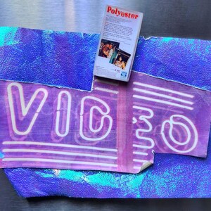 May include: A purple and blue abstract design with the word "VIDEO" in neon-style lettering. A VHS tape labeled "Polyester" rests on top. The background has a textured, iridescent appearance, creating a retro aesthetic.