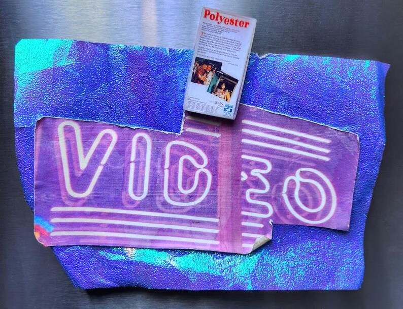 May include: A purple and blue abstract design with the word "VIDEO" in neon-style lettering. A VHS tape labeled "Polyester" rests on top. The background has a textured, iridescent appearance, creating a retro aesthetic.