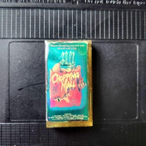 May include: A VHS tape of the movie "Chopping Mall" with a colorful cover featuring a monster and the text "Chopping Mall". The tape is set against a black background.
