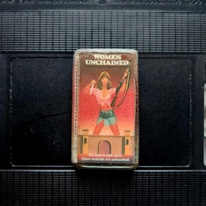 Könnte beinhalten: Eine Vintage-VHS-Kassette des Films "Women Unchained". Das Cover zeigt eine Frau mit Messer und Gewehr. Der Text auf dem Cover lautet "Women Unchained" und "No man is safe once these animals are unleashed."