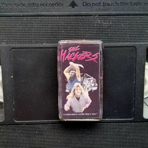 May include: A VHS video cassette of the film "The Hackers" with a vibrant cover design. The tape is placed on a black VCR cassette. The quote "A twisted mind is a terrible thing to waste" is displayed on the cover.