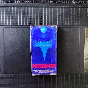 Puede incluir: Una cinta VHS azul de la película "Psycho Cop" con el texto "He's the last cop you'll ever meet." El título de la película es rosa y rojo. El fondo es negro.