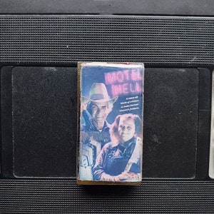 May include: A vintage VHS tape of the movie "Motel Hell" featuring a blue and white cover with two people. The text on the cover reads "Motel Hell" and "It takes all kinds of critters to make Farmer Vincent's fritters."
