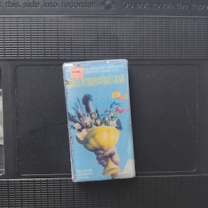 Może przedstawiać: Vintage kaseta VHS z filmu "Monty Python and the Holy Grail" z niebieską okładką przedstawiającą złoty kielich i ilustracje rycerzy. Taśma jest umieszczona na czarnym tle, z widocznym tekstem na grzbiecie.