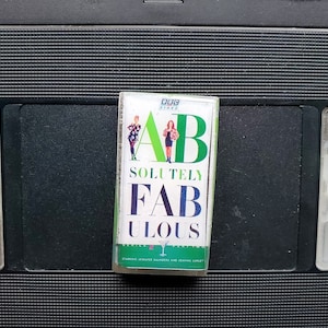 Könnte beinhalten: Eine VHS-Kassette der britischen Comedy-Serie Absolutely Fabulous mit dem Titel der Show in großen, farbigen Buchstaben. Die Kassette befindet sich vor einem schwarzen Hintergrund eines Videorekorders.