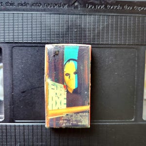May include: A small, rectangular VHS tape with a movie cover featuring a yellow-faced figure with a blue and green background. The tape is set against a black VHS cassette.