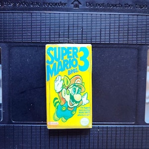 Puede incluir: Un cartucho de videojuego amarillo de Super Mario Bros. 3 con una imagen de dibujos animados de Mario. El cartucho está sobre un fondo de cinta VHS negra. El texto en el cartucho dice "SUPER MARIO 3 BROS."