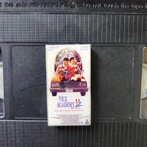 May include: A VHS tape of the film "Vice Academy 2" with a colourful cover showing actors in a car. The tape is set against a black background of a larger VHS cassette.