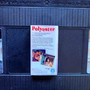 May include: A VHS tape labeled "Polyester" with a white label and red text. The label includes text and images from the movie. The tape is set against a black background of a VCR.