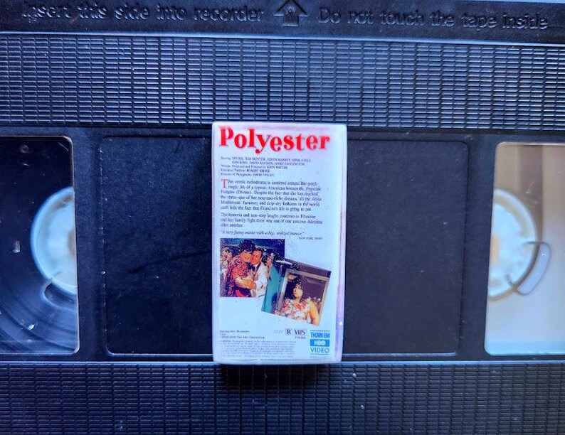 May include: A VHS tape labeled "Polyester" with a white label and red text. The label includes text and images from the movie. The tape is set against a black background of a VCR.