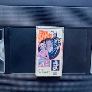 Op de afbeelding: Een vintage VHS-band voor de film "Evil Spawn" wordt getoond. Het label van de band toont een vrouw, een monster en een man. De tekst "Evil Spawn" is in een gouden, gestileerde lettertype. De band staat tegen een zwarte achtergrond.