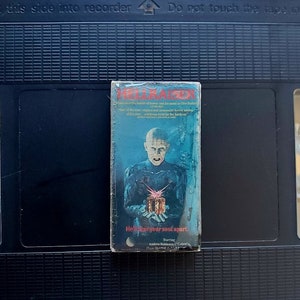 May include: A VHS tape of the horror film "Hellraiser" featuring a blue and black cover with a character holding a puzzle box. The text "Hear your soul apart" is visible. The tape is inside a black VHS case.