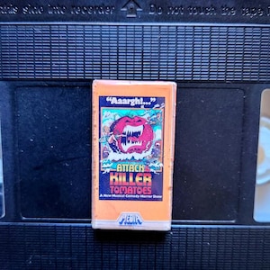 May include: A vintage VHS tape of "Attack of the Killer Tomatoes" with a colorful cover. The cover features a cartoon tomato with sharp teeth and the text "Aaargh!". The tape is set against a black background.