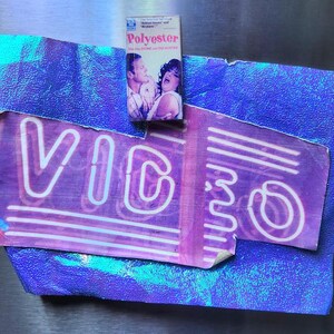May include: A vintage VHS tape labeled "Polyester" rests atop a torn purple and blue "VIDEO" sign. The sign has white neon-style lettering and is set against a textured, iridescent blue background. The tape features a movie still.