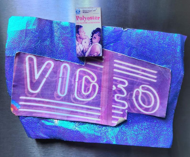May include: A vintage VHS tape labeled "Polyester" rests atop a torn purple and blue "VIDEO" sign. The sign has white neon-style lettering and is set against a textured, iridescent blue background. The tape features a movie still.
