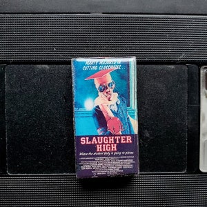 Peut inclure: Une cassette VHS avec le titre du film "Slaughter High" sur l'étiquette. L'étiquette présente une illustration colorée d'une personne portant des lunettes de soleil et un chapeau de remise de diplôme. La cassette est posée sur un fond noir.