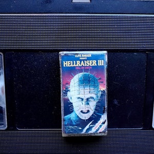 May include: A vintage VHS tape of the horror film "Hellraiser III: Hell on Earth" by Clive Barker. The cover features a blue and red graphic of the character Pinhead. The tape is set against a black background.