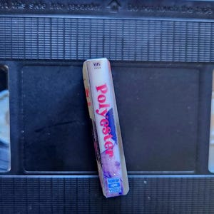 May include: A VHS tape labeled "Polyester" in red and white lettering, with a blue and purple design. The tape is resting on a black VHS cassette.