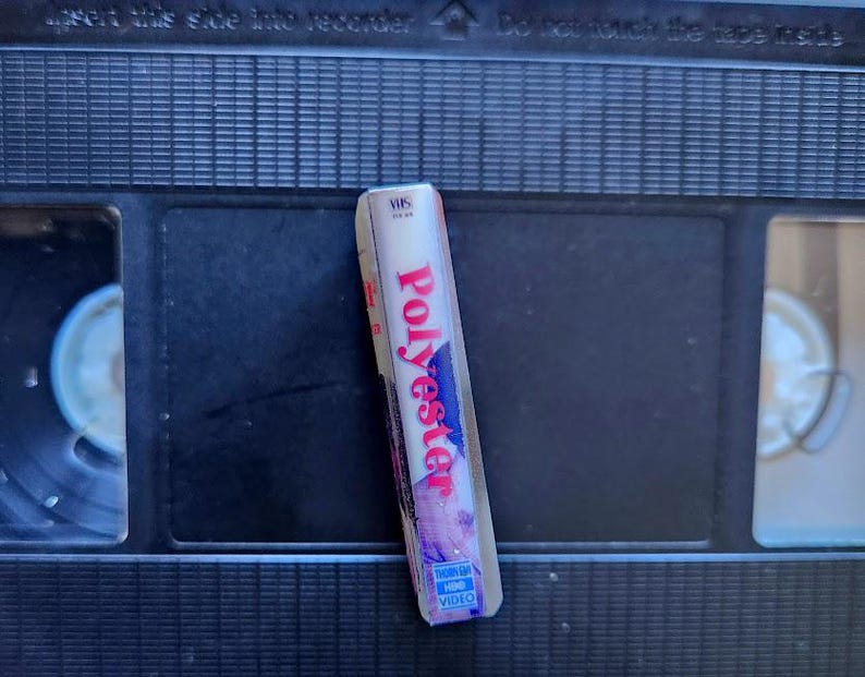 May include: A VHS tape labeled "Polyester" in red and white lettering, with a blue and purple design. The tape is resting on a black VHS cassette.