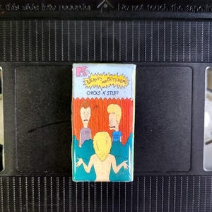 Può includere: Un magnete rettangolare a forma di cassetta VHS di Beavis and Butt-Head. Il magnete presenta il logo MTV e il testo "BEAVIS and BUTTHEAD CHICKS N' STUFF". L'opera d'arte raffigura i due personaggi e una figura bionda.