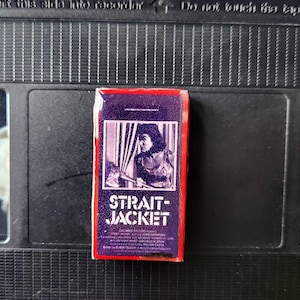 May include: A small, rectangular VHS tape box with a red border and the title "STRAIT-JACKET" in white letters. The box features a black and white image of a person holding an axe. The box is resting on a black VHS tape.