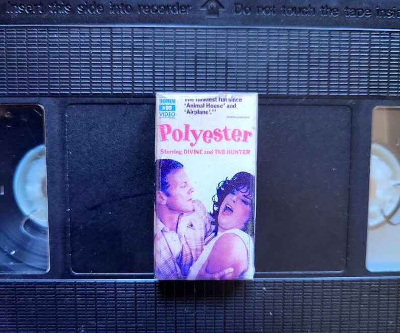 May include: A VHS tape for the movie "Polyester" is displayed. The tape's cover features a colorful design with the movie title and the actors' names. The tape is set against a black background.