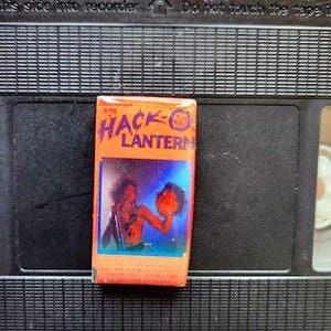 May include: An orange VHS tape box with the title "The Hack Lantern" in black and yellow lettering. The box features a colourful illustration of a person holding a glowing object. The tape is set against a black background.