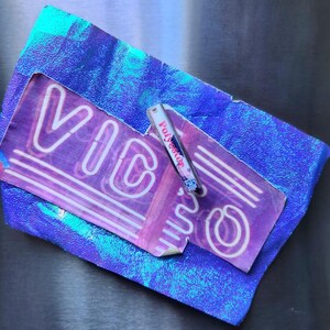 May include: A purple and white neon sign with the word "VIC" is displayed on a textured surface. A small cassette tape labeled "Polyester" rests on the sign. The background is a shiny, iridescent blue.