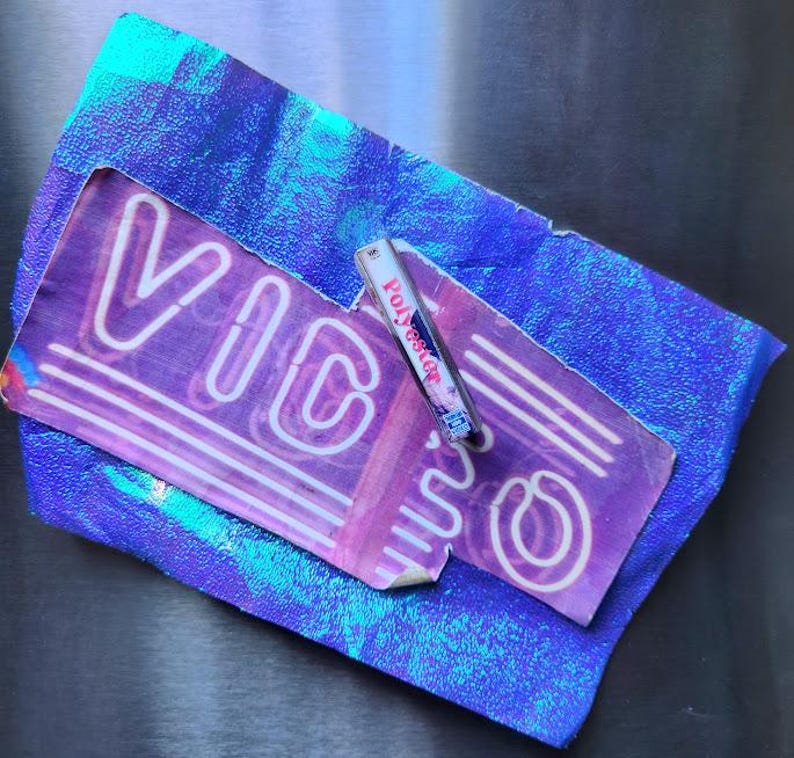 May include: A purple and white neon sign with the word "VIC" is displayed on a textured surface. A small cassette tape labeled "Polyester" rests on the sign. The background is a shiny, iridescent blue.