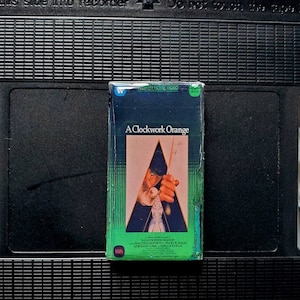 May include: A VHS tape of the film "A Clockwork Orange" with a green and blue cover featuring a central image of a person holding a weapon. The tape is inside a black VCR cassette.