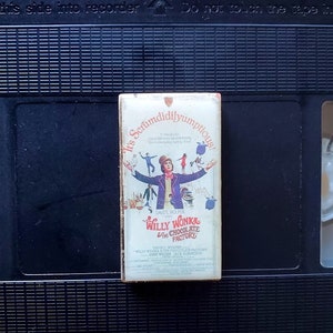 May include: A vintage VHS tape of "Willy Wonka & the Chocolate Factory" with a colorful movie poster design. The tape is in a black VHS player. The movie title and "It's Scrumdidilyumptious!" are visible on the tape.