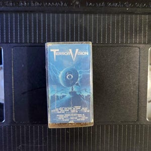 May include: A VHS tape for the film "TerrorVision" with a blue and white cover. The cover art depicts an eye-like image with lightning. The tape is set against a black background, with the VHS case in the centre.