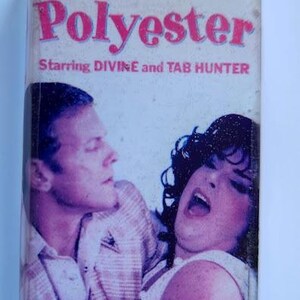 May include: A VHS tape for the movie "Polyester" starring Divine and Tab Hunter. The cover features a pink title and images of the actors. The HBO Video logo is visible in the upper left corner.