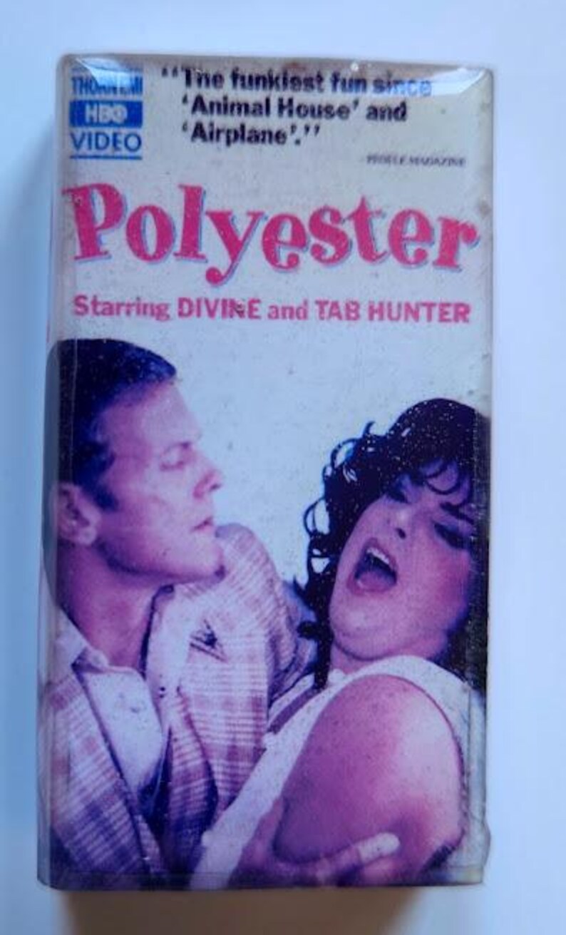 May include: A VHS tape for the movie "Polyester" starring Divine and Tab Hunter. The cover features a pink title and images of the actors. The HBO Video logo is visible in the upper left corner.