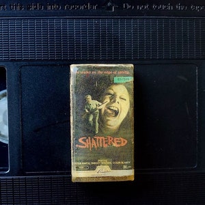 Puede incluir: Una cinta VHS vintage de la película "Shattered" con una cubierta deteriorada que muestra a una mujer gritando y a un hombre. La cinta está colocada sobre un reproductor VHS negro.