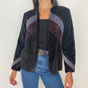 May include: Black quilted velvet jacket with gray and burgundy accents. The jacket has a stand-up collar and a vertical stripe design. The jacket is open in the front and has long sleeves.